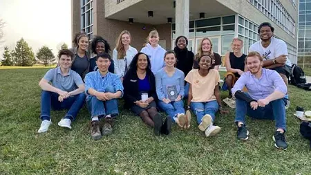 Group of 13 students and faculty member smiling outside, with some holding a book.